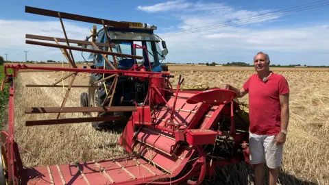 A red binding machine for harvesting long straw. It has a high cylindrical cutting blade which gathers the straw on to a red conveyor belt system, which the gathers and ties the straw into bundles