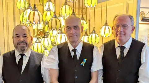 Empire Hotel Jean Paul, Nelson and David all wearing white shirts, black ties, black waistcoats  and white aprons standing in front of a cluster of bright pendant lights