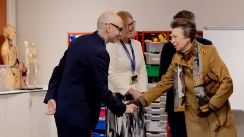 Princess Anne shakes hands with a male staff member in a medical learning room at the university campus, other staff members are stood around them