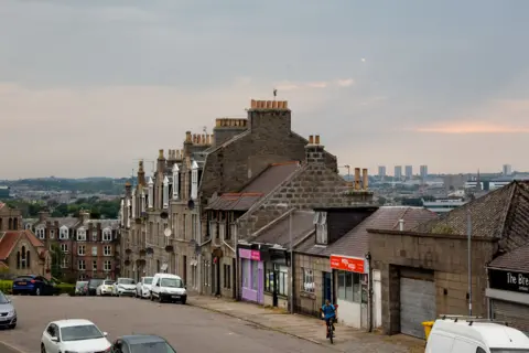 Getty Images é uma imagem de rua no Tory de Aberdin. Vemos uma fileira de lojas com apartamentos e alturas terelas misturados. O fundo contém montanhas e apartamentos altos.