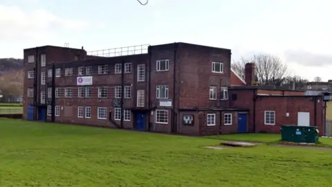 Picture shows a 1930s style red brick building with three floors, lots of windows and a flat roof, sitting in the middle of a field with fences surrounding it.