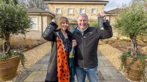 Omaze A man and a woman standing in front of a six bedroom house in Somerset, England. Both smiling, the man is holding keys and the woman is holding a black bottle of alcohol