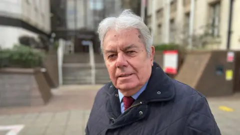 Mike Mackrory, a man in his 70s, is smiling at the camera with an out of focus council building behind him.