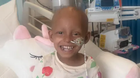 Family photograph Ariyah is sat in a hospital bed smiling. She has a pony cushion surrounding her. She has very little hair on her head and and has a thin tube up her nose. she is wearing pyjamas with red cherries printed on them. 