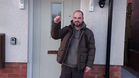 A man with short cropped brown hair and a brown beard. He is wearing a brown puffer jacket and holding up house keys outside a new home. 