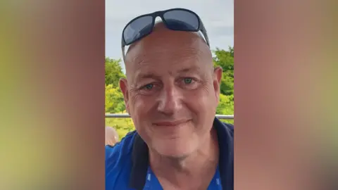Jeff Blair with sunglasses on his head and wearing a blue top. He is bald and is smiling in the close-up shot. There are trees behind him.