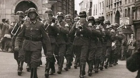 German soldiers in uniform, some playing musical instruments, march in front of the Lloyds bank building in St Peter Port
