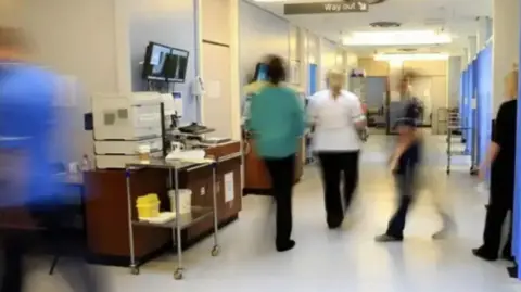 A generic image of medical staff, who have been blurred visually, moving around a hospital attending to patients. They are dressed in various types of medical scrubs and can be seen walking through a ward which has patient areas on the right-hand side.