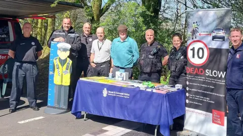 Devon and Cornwall Police Seven men and one woman stand in a row smiling. Three of them are wearing police uniform and two are wearing fire service uniforms. Some are standing behind a table with a Devon and Cornwall Police branded tablecloth which is covered with leaflets. Another has a cardboard cut out of a cartoon police officer with a ruler along its side. There is a British Horse Society pop-up banner that reads: 10 DEAD SLOW. It has a photo of a car measured to be two metres away from two horse riders.