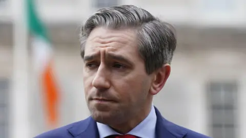 PA Media Harris has short grey and black hair, dark eyebrows and short facial hair. He has a navy blazer, a red tie and a white shirt. The background behind him is blurred.