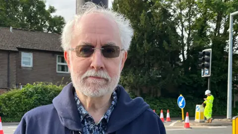 An older man with white hair and sunglasses stood at a roadside looking into the camera with a blank .