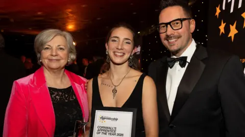 Truro and Penwith College A young woman receives an award certificate, smiling for the camera. She is flanked by an older woman wearing a raspberry coloured velvet jacket and a man with glasses wearing a dinner jacket 