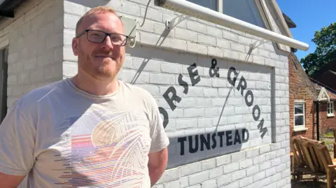 Owen Sennitt/BBC A man with glasses and a short beard smiles at the camera in front of a pub building which is painted white, with the Horse & Groom logo painted on
