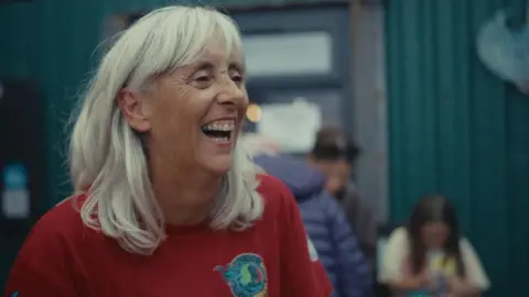 Braving the Blue - Our Lives, BBC Cymru Wales Makala, who has white straight hair and wears a red t-shirt, smiling at a fundraising event. 