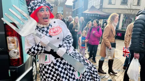 The image shows a costumed performer dressed in a black‑and‑white checkered outfit, decorated with food-themed patches. The costume includes a matching checkered hat with bright red hair attached, a white top with large buttons, and a badge that reads I Love Rhubarb.