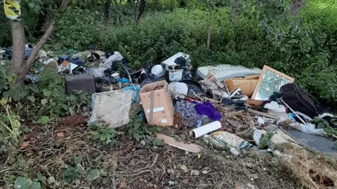 North Northamptonshire Council A pile of fly-tipped rubbish spread across the edge of a verge. There are boxes, rolls of foam, a mattress, building waste and black plastic bags of waste. Behind it are trees and nettles. 