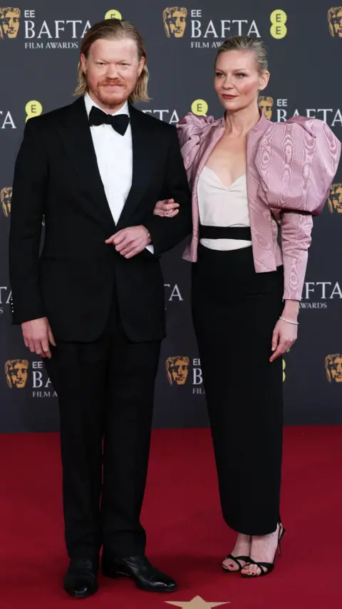 Getty Images Jesse Plemons and Kirsten Dunst - he is in black tie and she is in a pink jacket and black trousers