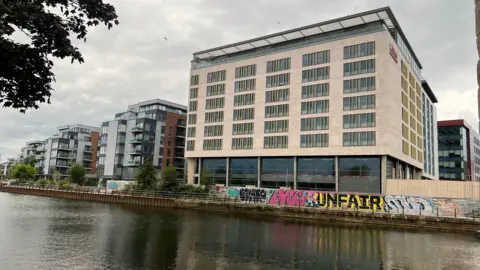 View of the Hilton Garden Inn building from the river. There is graffiti on a wall in front of the hotel. 