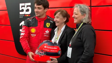 Charles Leclerc posing with Joann Villeneuve and Melanie while holding his helmet that pays tribute to Gilles Villeneuve