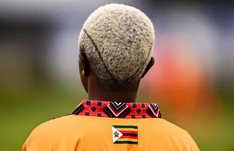BEN MCSHANE / SPORTSFILE / GETTY IMAGES A woman with cropped hair that is dyed blonde stands with her back to the camera. She wears the Zimbabwe kit, which is bight orange with a small Zimbabwean flag at the nape of the neck.