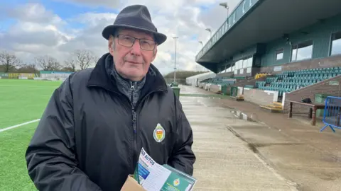 Juliette Parkin / BBC A man wearing a black coat and hat in a football stadium holding a cardboard box of football programmes.