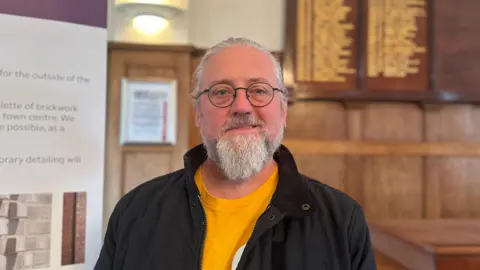 A man with tied back white hair, a white beard an round glasses smiles at the camera. He is wearing a black jacket and orange t-shirt.