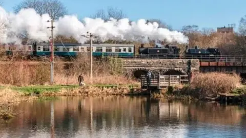 A green heritage train passing over a viaduct above a river on a sunny day with steam billowing overhead as man walks his dog along a river bank under the structure.