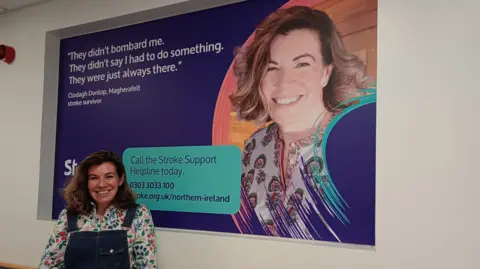 Stroke Association. Shows a woman in denim dungarees in front of a poster with her on it.