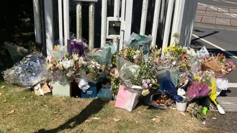 Floral tributes left by the crossing on a patch of grass with a white railing behind