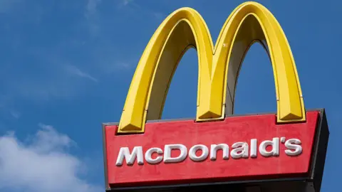 The McDonald's golden arches sign with a large yellow letter M above the name McDonald's written in white on a red background. There is a blue sky behind it.