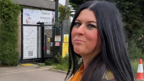 A woman with long black hair standing outside near a green hedge, looks sideways towards the camera