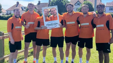 Freddie Pring Memorial Fund A group of men is wearing orange football kit and is standing arm in arm on grass.