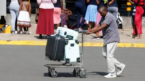 A side view of a man in trousers and a T-shirt wheeling a luggage trolley with suitcases along a road. Other airport users can be seen behind him.