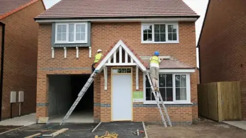 Getty Images Two men wearing hi-vis vests standing up ladders either side of the doorway of a new build house.