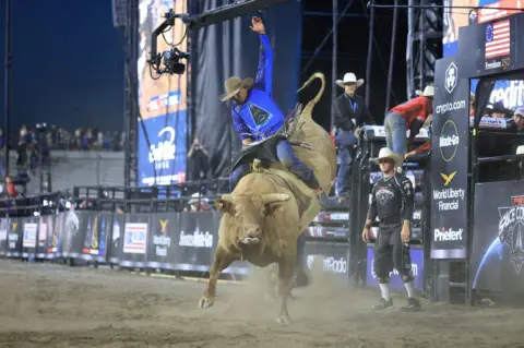A professional bull rider, wearing a cowboy hat and blue shirt featuring the triangular logo of US Space Force, clings on while riding on the back of a massive, brown horned bull in a stadium filled with dirt 