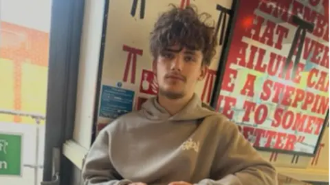 Family handout Alfie Moran, wearing a cream-coloured hoodie, sits on a stool holding his phone. He has long brown hair swept down in a fringe. 