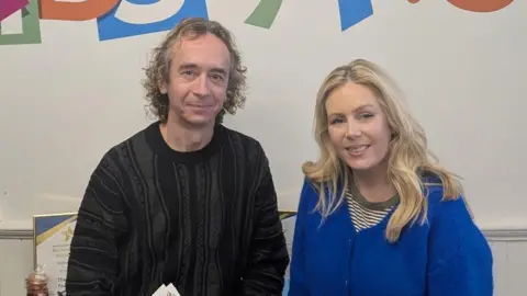 Geoff Russell-Jones in a black jumper with curly hair smiles at the camera while standing next to Rebecca Caswell-Fox, who has long blonde hair and a blue cardigan.