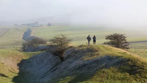 Getty Images A couple and their dog walk along earthworks in the North Wessex Downs. There are fields for as far as the eye can see surrounding the raised ground but morning mist is obscuring the view.