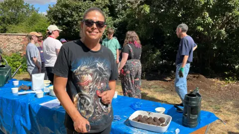 Participant Jasmin making wildflower seed balls in the grounds of the cathedral. She is wearing a black t-shirt with a dragon on it and dark sunglasses, and other volunteers can be seen behind her.