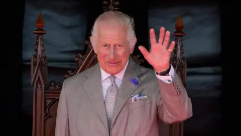 A picture of King Charles. He is waving and wearing a grey suit with blue accents and is stood in front of a wooden chair. 