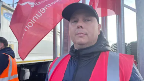 A man standing in front of a large metal fence with a red unite the union flag flowing in the wind. He is wearing a black hat and black coat with a pink unite hi-vis jacket on top