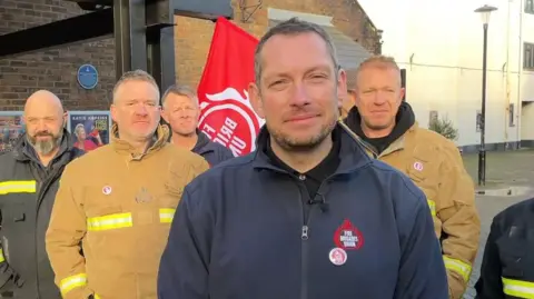Image of a group of firefighters standing with banners and flags outside a red brick building.