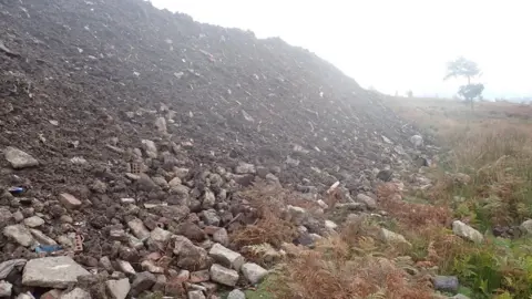 A large pile of rubble and dirt has been dumped in the middle of a grassy field. A tree can be seen in the distance.