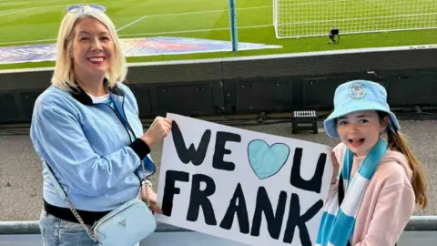 Handout A woman and her daughter stood in the crowd infront of a football pitch holding a sign that says we love you frank. 