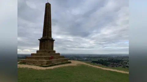 A view from behind the War Memorial, looking out over the landscape below Ham Hill. The memorial itself consists of four steps with a large stone obelisk in the middle. The names of the dead are written on tablets on its side.