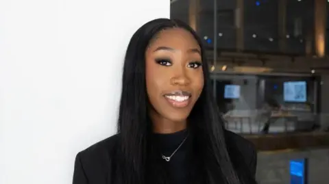 Handout Renee Washington wears a black t-shirt and has long black hair. She is standing in front of a white wall and a glass wall. 
