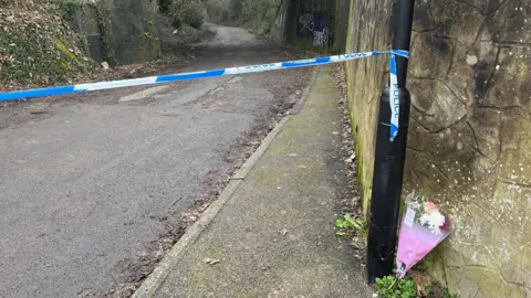 A blue and white police cordon is attached to a lamp post. Behind it there is a path surrounded by trees that goes underneath a railway bridge. There is a bunch of flowers in pink cellophane wrapping resting against the wall next to the lap post.