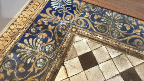 Close up photograph of ornate patterned floor tiles. Mosaic stiles borders in blue and gold with a diamond pattered white and black centre.
