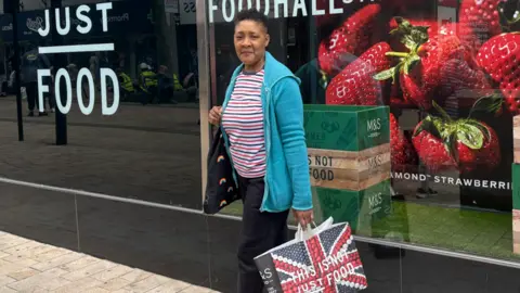 Woman looking into camera while standing outside M&S shop with an M&S bag in her hand. 