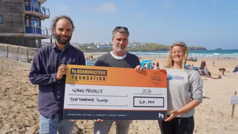 Two men and a woman hold a large novelty cheque and smile at the camera. They are standing on a sandy beach with the sea in the background. The cheque is made out to the Wave Project for £10,000.